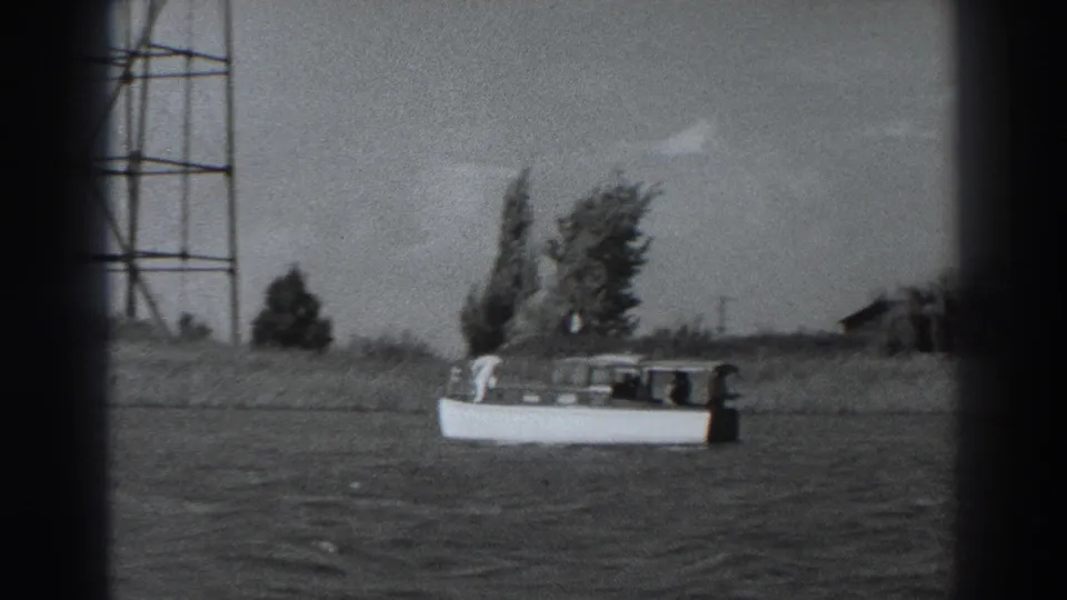 1938 Fort Ross home-movie frame showing a small white motorboat offshore, with trees and a tall metal tower along the background shoreline.