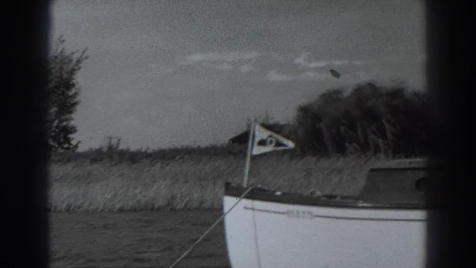 1938 Fort Ross home-movie frame showing a small white boat at the edge of a grassy shoreline with reeds, brush, and an overcast sky.
