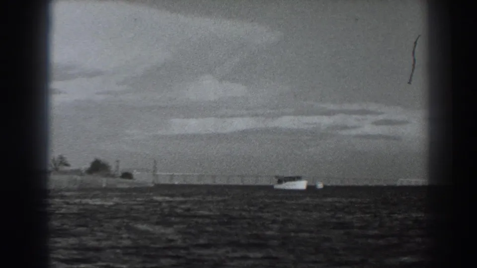 Black-and-white 1938 Fort Ross frame showing a small boat on rippled coastal water, with a low shore and overcast sky in the distance.
