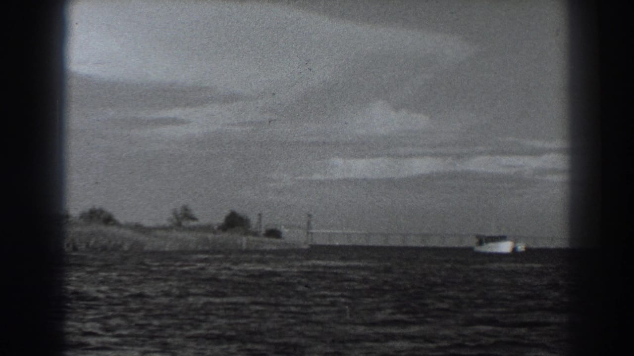 1938 8mm frame near Fort Ross showing open water, a low shoreline, cloudy sky, and a small distant vessel.
