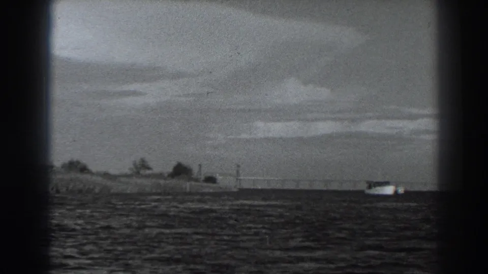 1938 8mm frame near Fort Ross showing open water, a low shoreline, cloudy sky, and a small distant vessel.