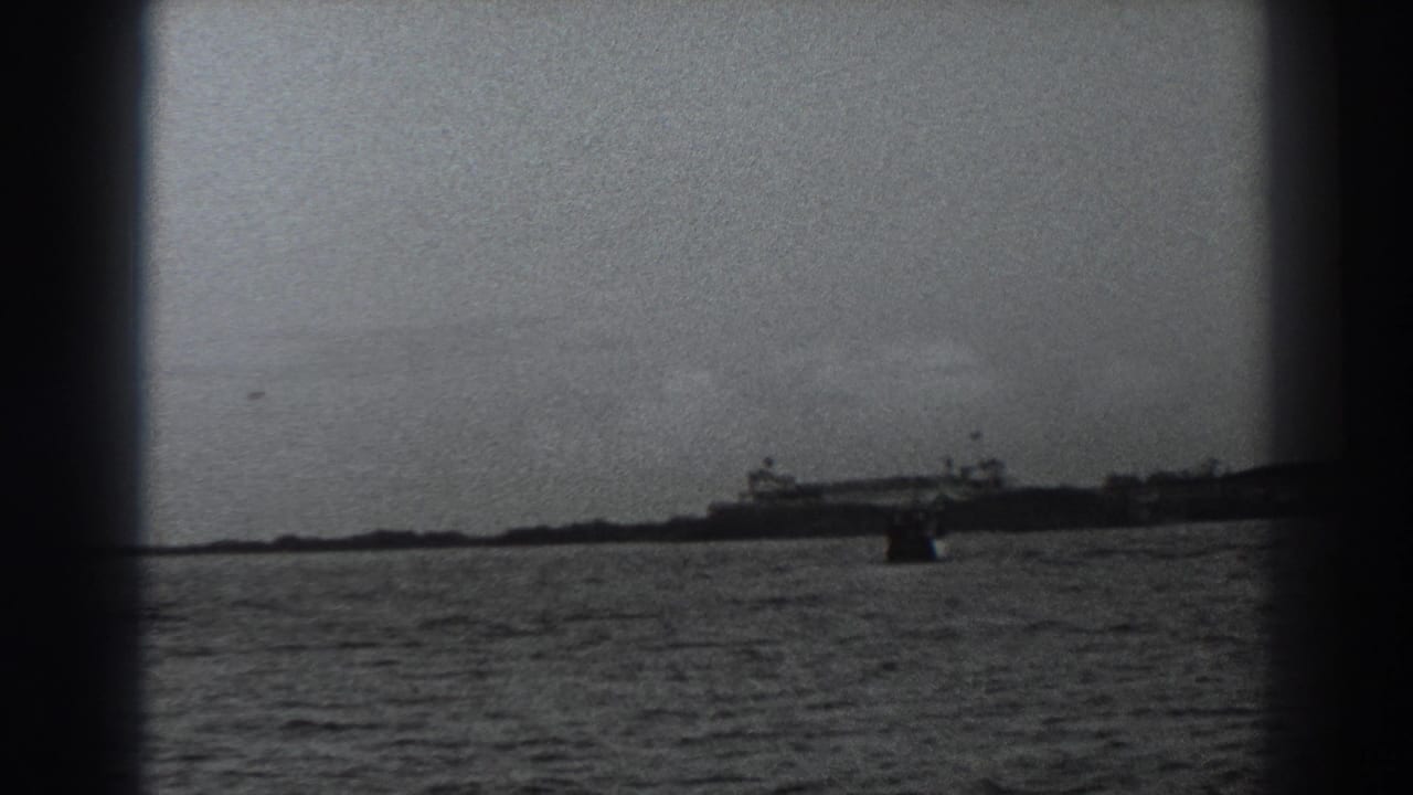 Distant shoreline and buildings across choppy water in a grainy black-and-white frame.