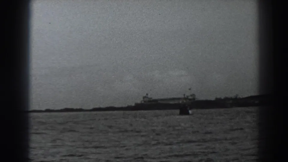Black-and-white 8mm view toward the Fort Ross-area shore, with distant buildings and a small boat-like shape on the water.