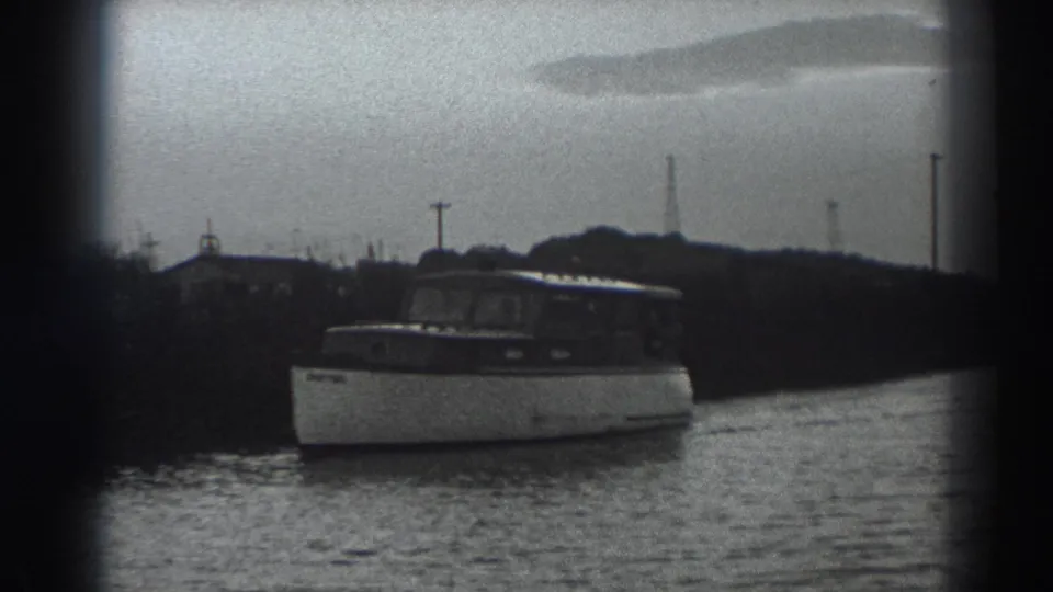 1938 Fort Ross home-movie frame showing a small cabin motorboat moving near shore with utility poles and low buildings in the distance.