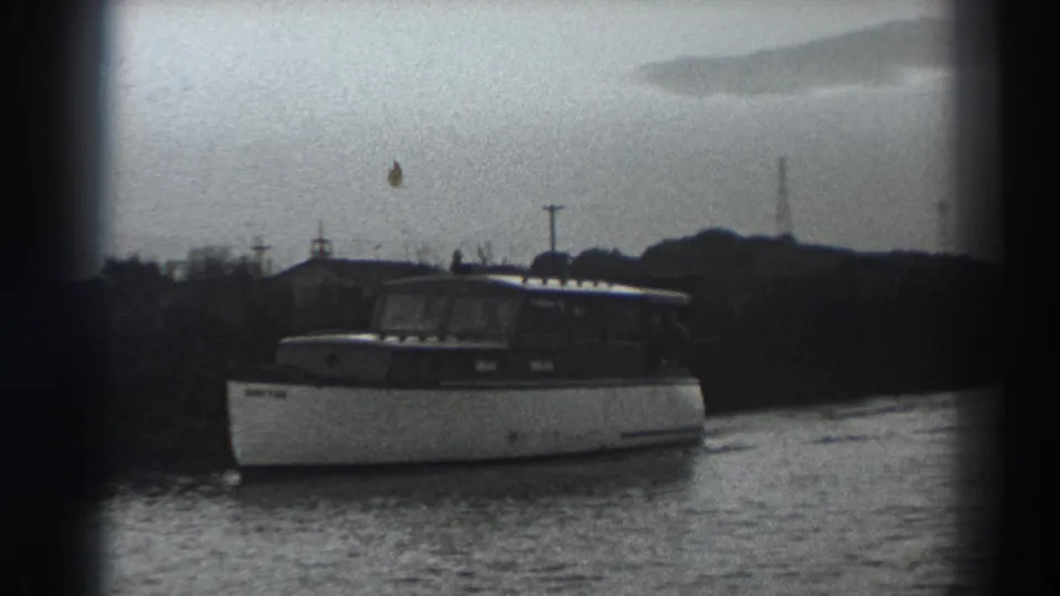 The Fort Ross boat remains centered on the water, with an overcast sky, low shoreline growth and distant poles visible behind it.