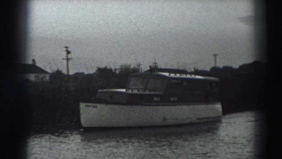 A closer 8mm view shows the cabin motorboat beside the shore, with rooflines, poles and soft black-and-white grain in the background.