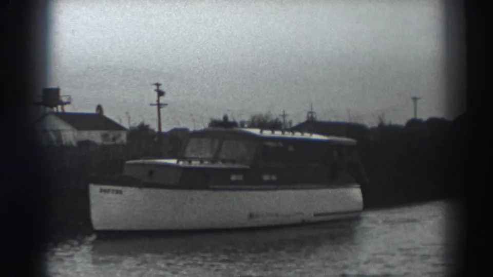 The boat moves along the Fort Ross shoreline, with small buildings, poles and a tower-like structure visible through soft 8mm grain.