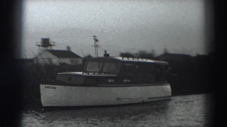 1938 Fort Ross-area 8mm frame showing a cabin motorboat moving past a low riverbank with small structures in the background.