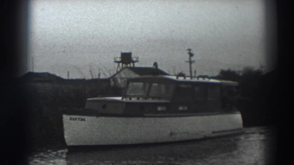 The motorboat continues along the Fort Ross-area riverbank, with a small tower-like structure and low buildings beyond the water.