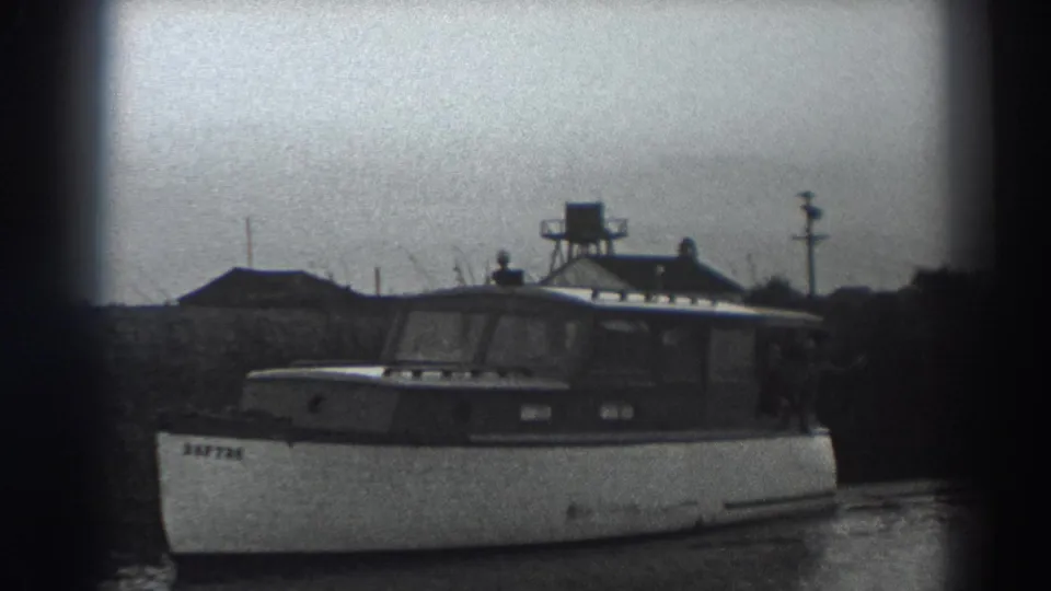 A closer 1938 8mm view of the cabin motorboat as it passes shoreline brush and low riverside structures near Fort Ross, California.