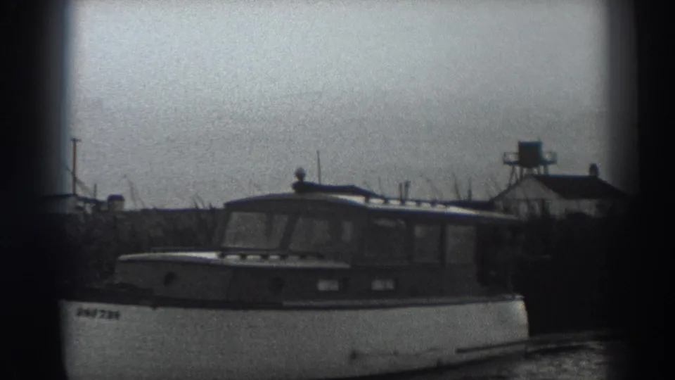 1938 Fort Ross home-movie frame showing a cabin motorboat passing near low shoreline buildings under an overcast sky.