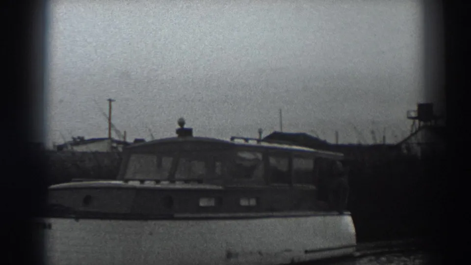 The cabin motorboat continues past the Fort Ross shoreline, with small buildings and a water tower visible behind it.