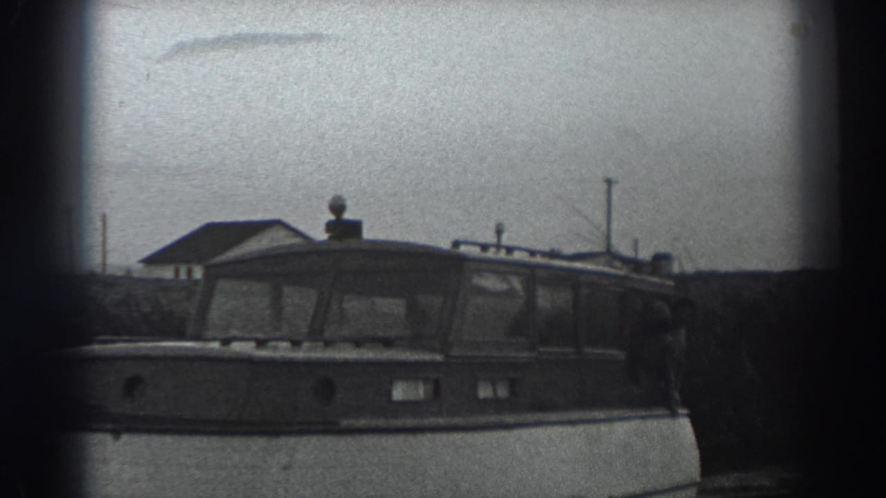 Cabin motorboat on calm water with trees and a roofline behind it.