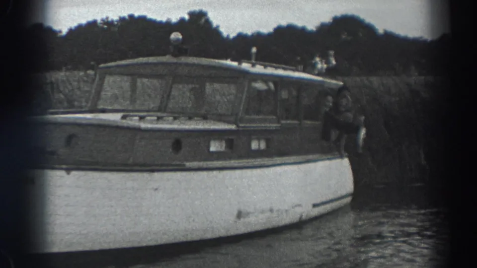 Black-and-white Fort Ross frame of a moored wooden cabin boat, with a person visible at the stern and trees beyond the bank.