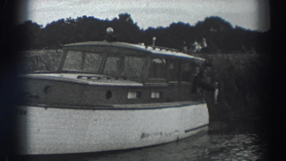 1938 black-and-white home-movie frame showing the side of a wooden motorboat near shore, with people partly visible behind the cabin.