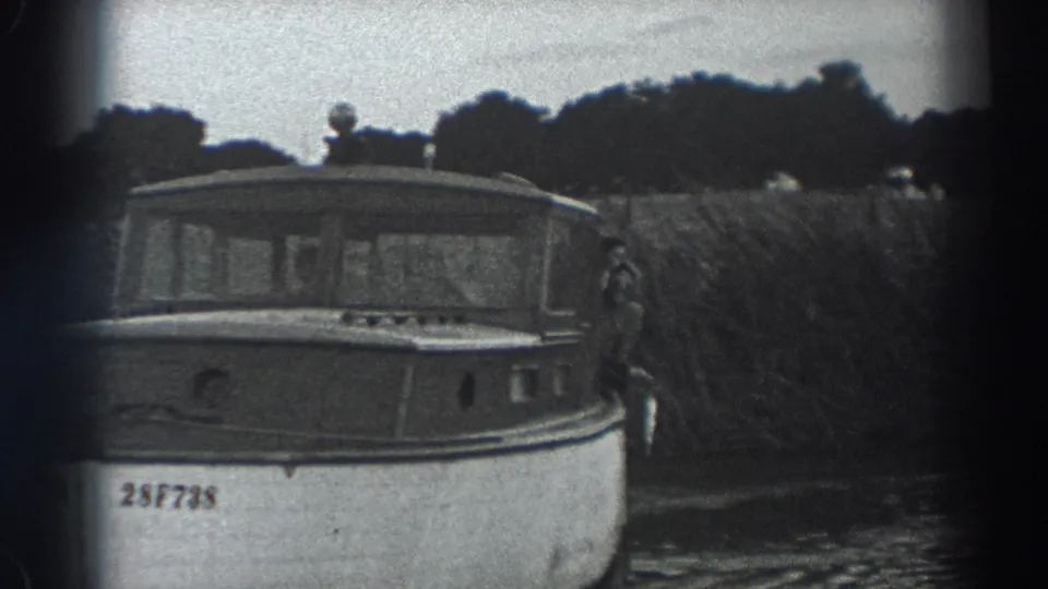 Black-and-white 8mm frame from Fort Ross in 1938 showing the cabin section of a small boat near a reed-lined bank.