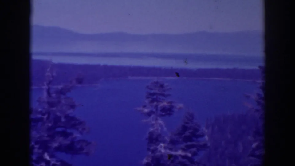 1938 Lake Tahoe-area 8mm vista showing blue water, foreground evergreens, haze, and low mountains in the distance.