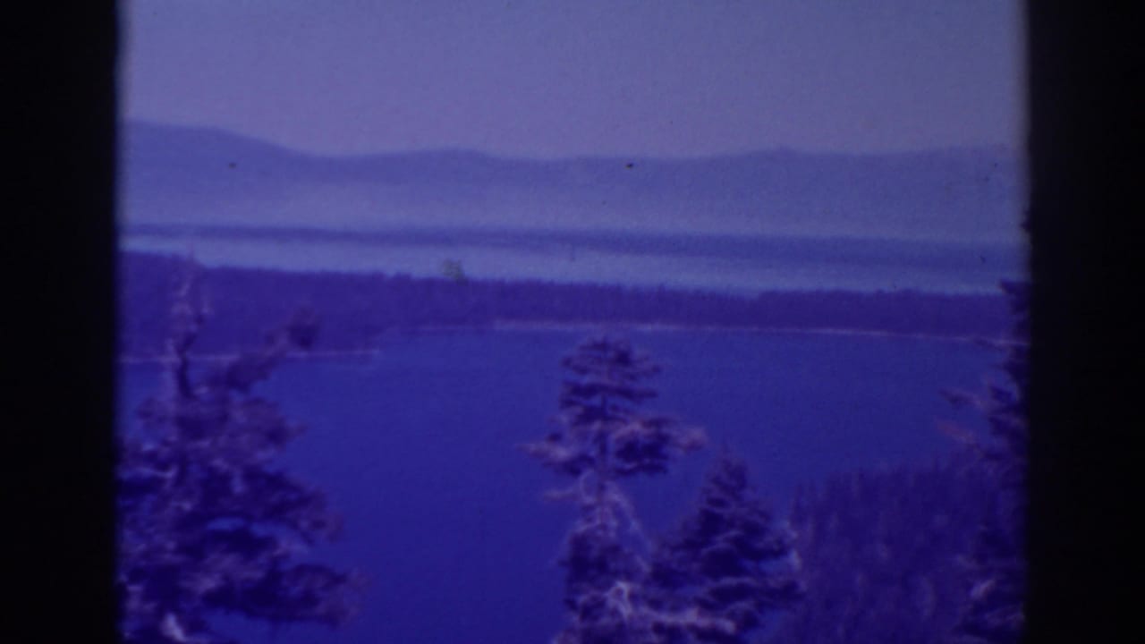 Blue-tinted lake view framed by dark trees with distant mountains beyond.