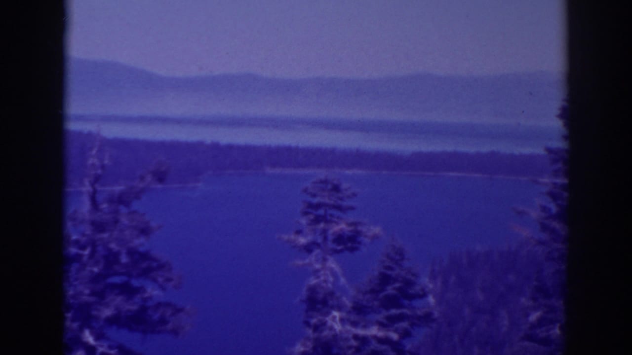 Lake and distant mountain ridges seen through dark tree forms.