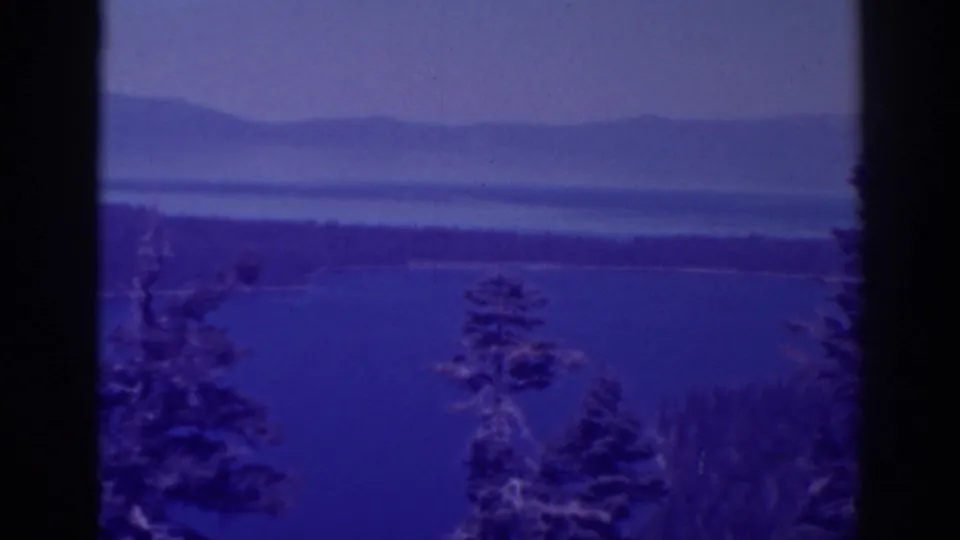 Late-1930s Lake Tahoe frame with tree silhouettes, lake water, a low shoreline, and hazy mountain ridges beyond.