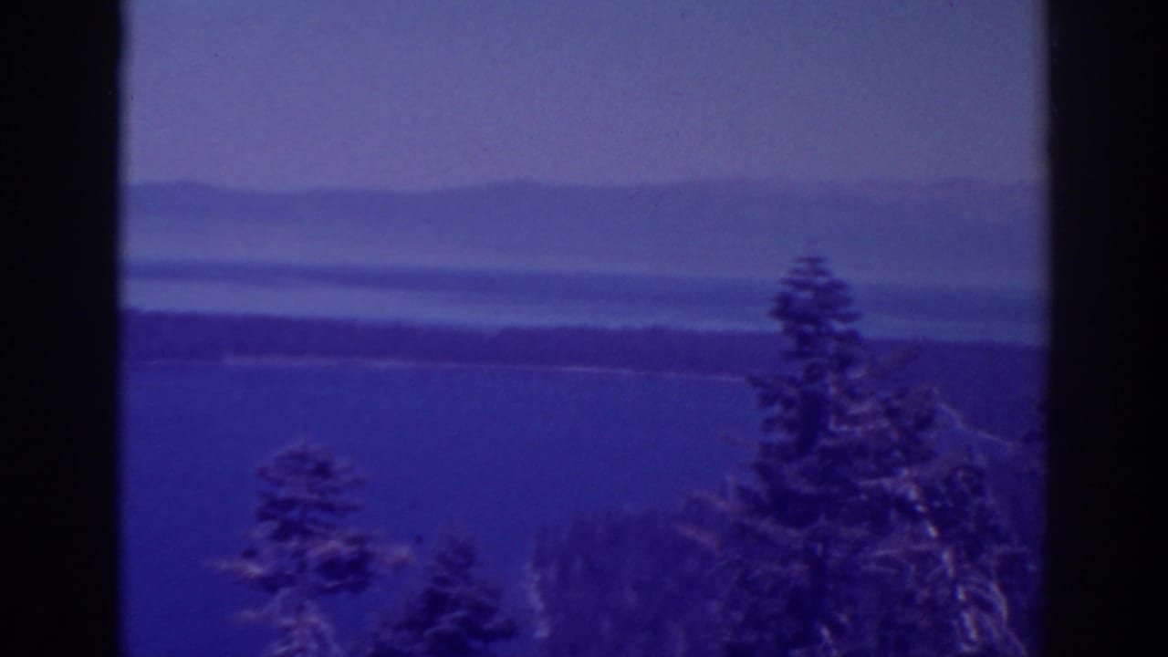 1938 8mm Lake Tahoe view with evergreen trees in the foreground, blue water, distant shoreline, and hazy hills under a pale sky.