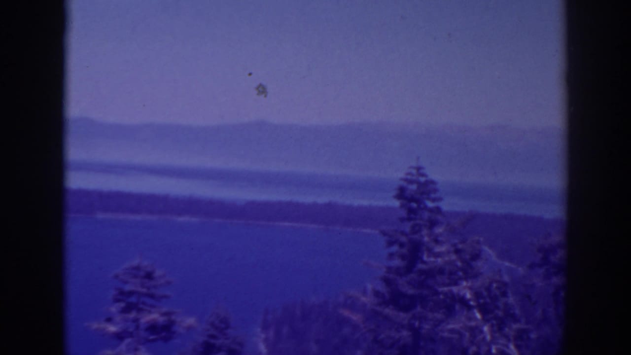 1938 Lake Tahoe home-movie frame with evergreens in the foreground, blue water, a dark shoreline, and hazy mountain ridges beyond.