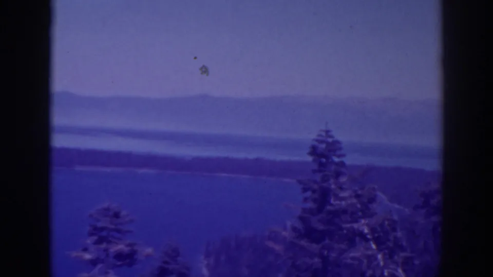 1938 Lake Tahoe home-movie frame with evergreens in the foreground, blue water, a dark shoreline, and hazy mountain ridges beyond.