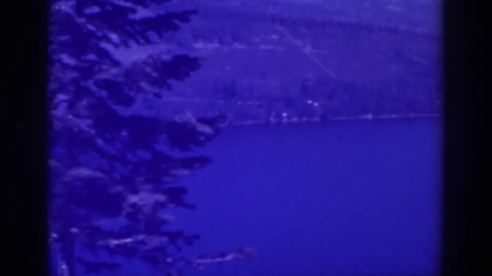 A closer 1938 Lake Tahoe frame shows dark evergreen branches over blue water, with a faint opposite shoreline and hillside beyond.
