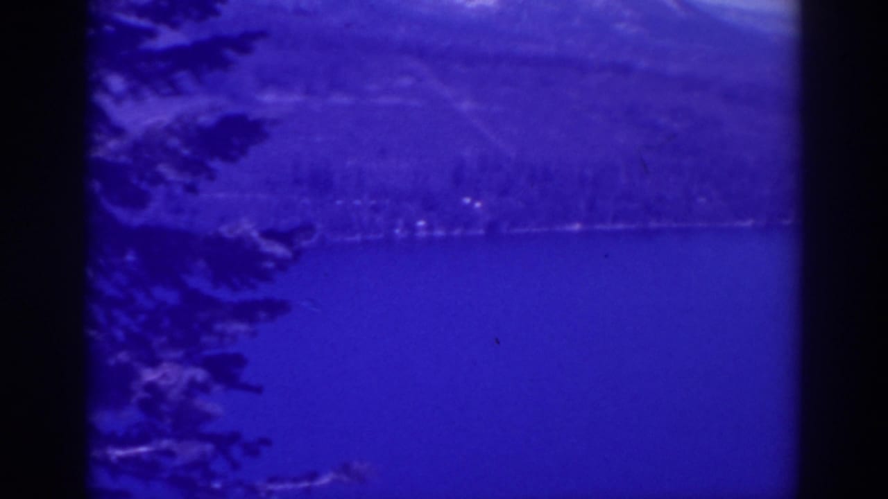 Blue-tinted lake with evergreen branches at left and distant mountains across the water.