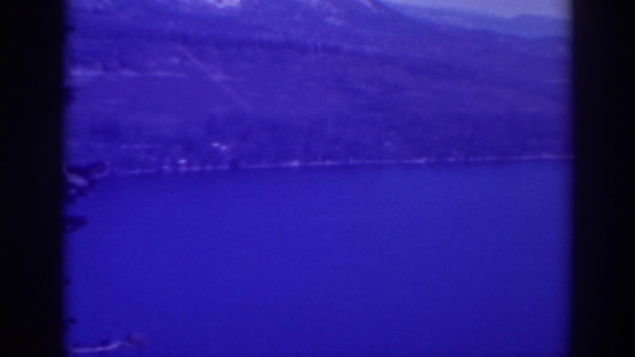 Calm blue lake beneath wooded hills and mountain ridges, partly framed by dark film edges.