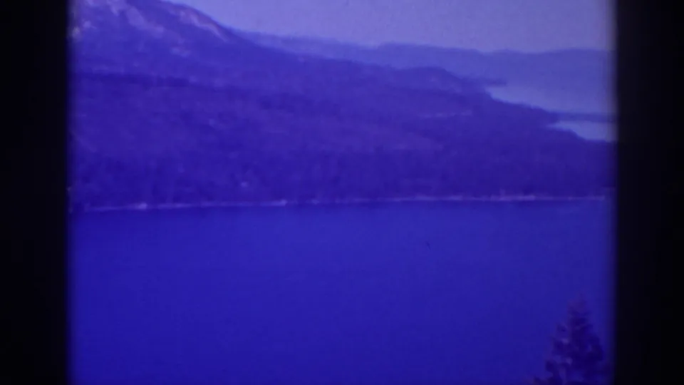 Late-frame Lake Tahoe view from 1938 home-movie footage, showing blue water, distant mountains, and a foreground tree edge.