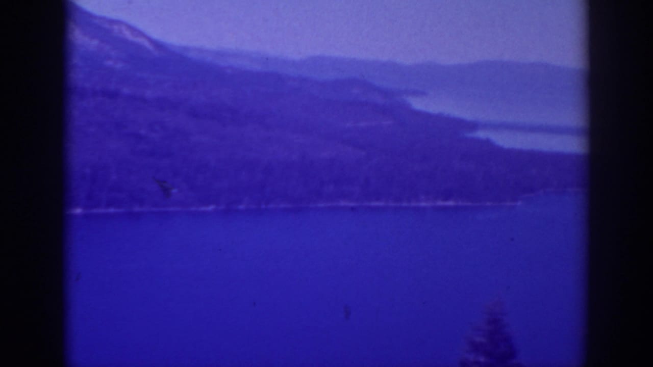 Blue-tinted lake and distant forested mountains seen through dark film-frame edges.