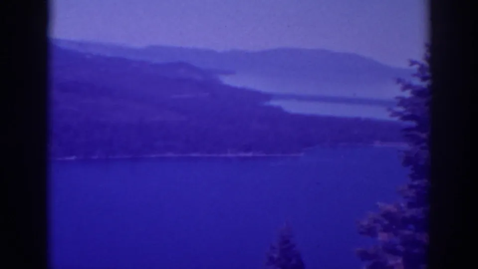 1938 Lake Tahoe frame with calm water below hazy ridges, bordered by dark film edges and foreground trees.