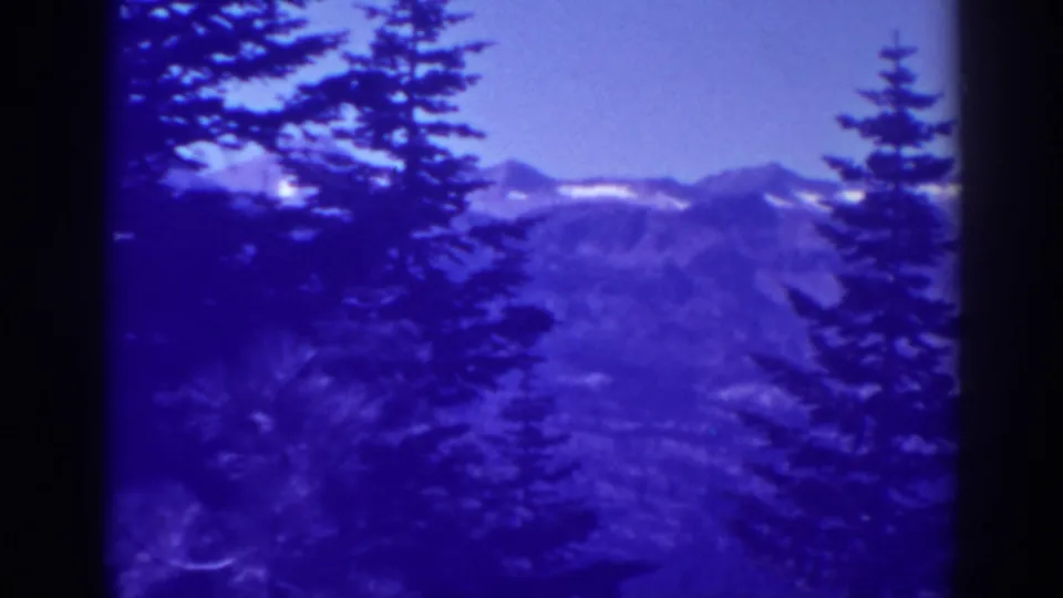 1938 Lake Tahoe home-movie frame showing dark evergreens before distant snow-streaked Sierra Nevada mountains.