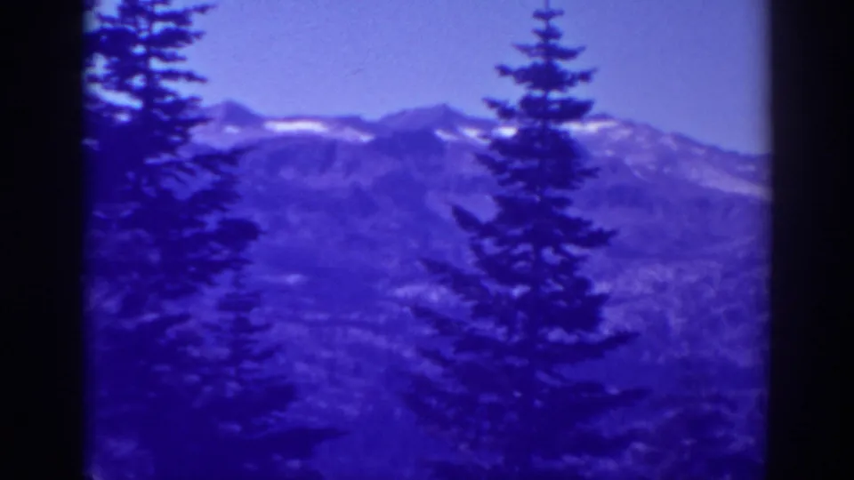Blue-tinted 8mm frame of Lake Tahoe forest and distant snow-streaked mountains, with no people visible.