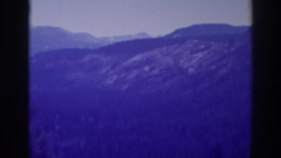 1938 Lake Tahoe 8mm frame of a blue-purple wooded slope, distant mountain layers, cloudy sky, and soft film texture.