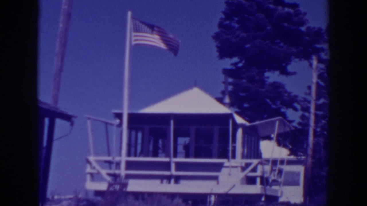 White lodge with porch, flagpole, American flag, blue sky, and pine trees.