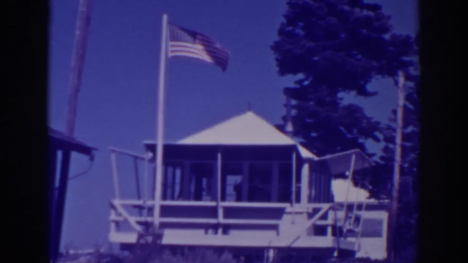 1938 Lake Tahoe home-movie frame showing a small white lodge, porch railings, American flag, clear sky, and tall pines.