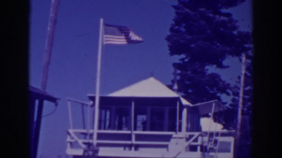 1938 Lake Tahoe frame with the flagpole centered near the white lodge roof, porch shadows, railings, and pine trees.