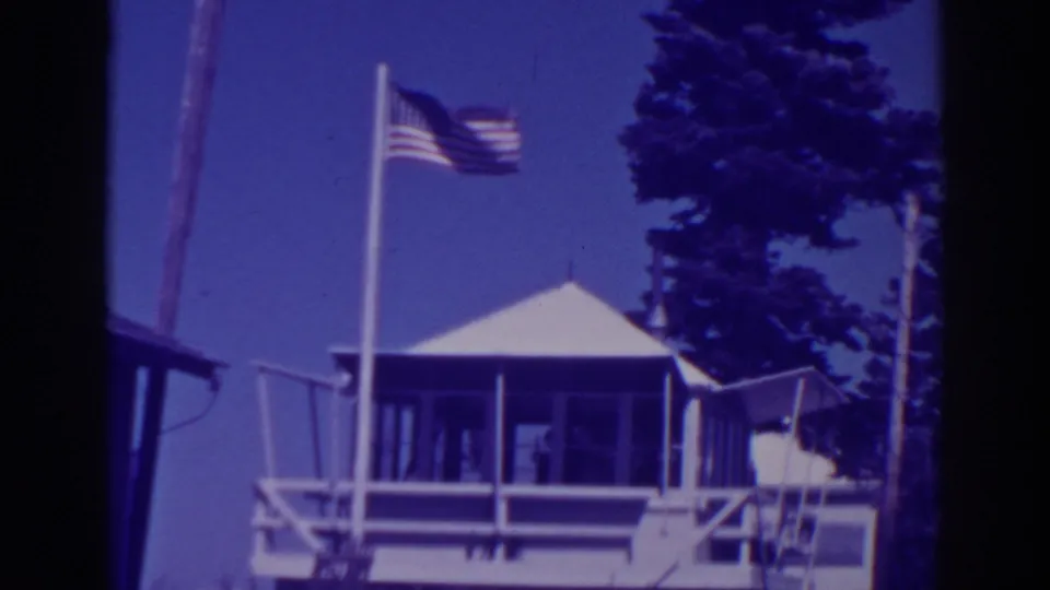 Late shot of the 1938 Lake Tahoe lodge exterior, showing the American flag, white porch structure, peaked roof, and pines.