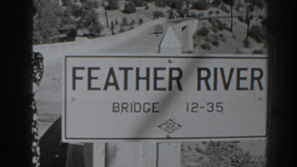 1938 Oroville footage frame showing the Feather River Bridge sign, bridge approach, distant road, and rural hillside.