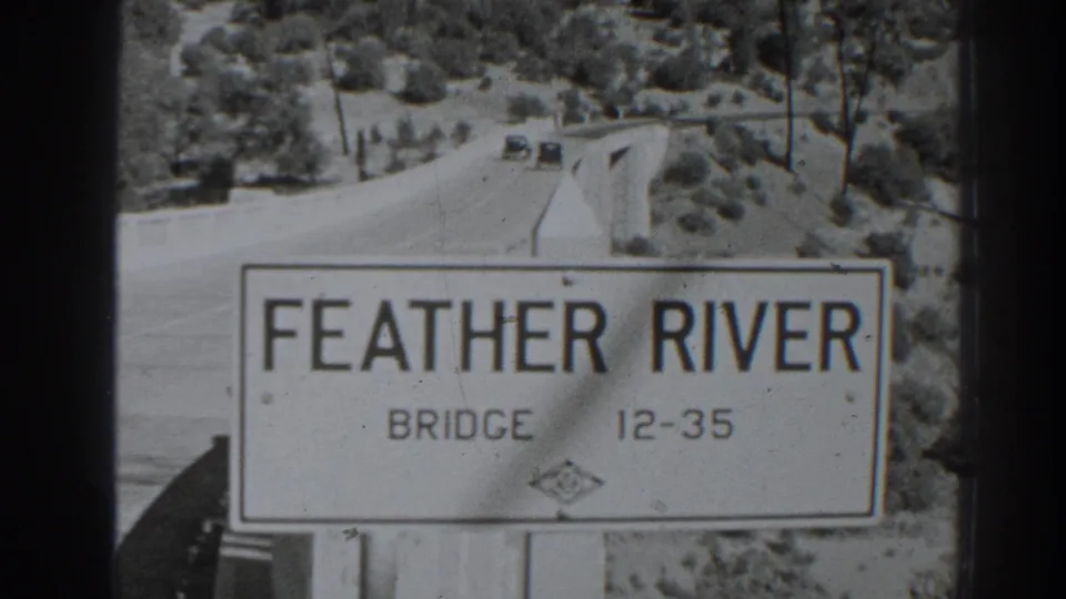 Black-and-white 8mm frame of the Feather River Bridge sign near Oroville, with the road and distant automobiles behind it.