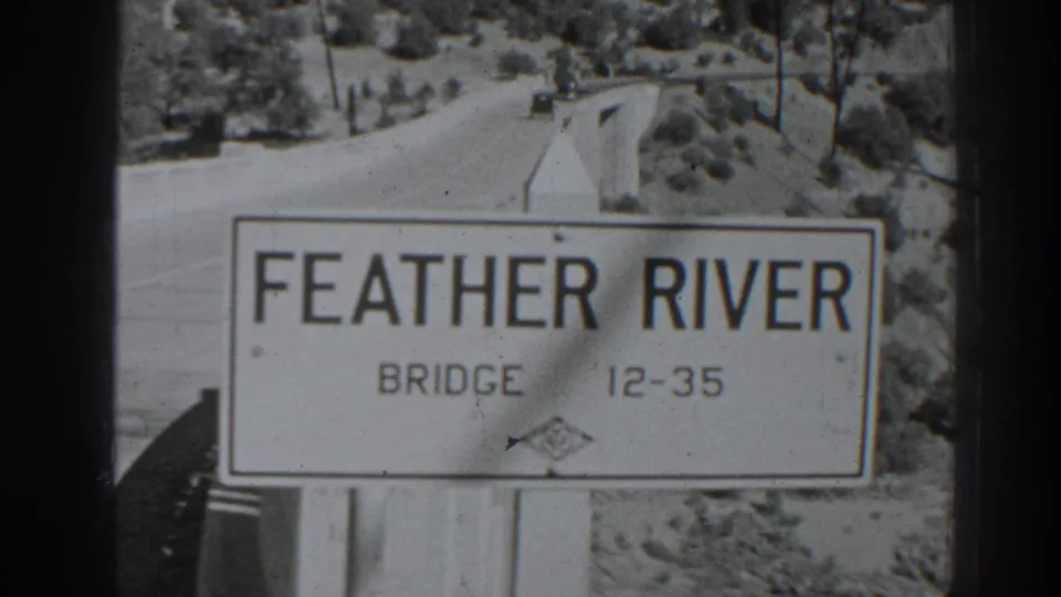 1938 8mm frame showing the Feather River Bridge sign in front of a rural road and tree-covered slope.