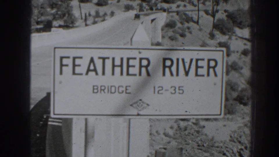 Late-1930s Oroville frame of the Feather River Bridge sign, with a pale roadway and rural hillside visible behind it.