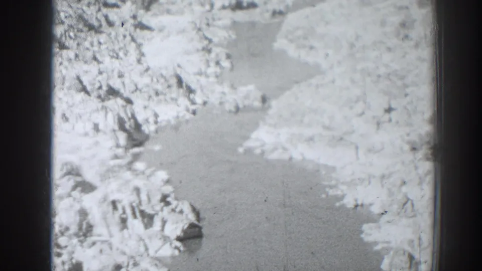 Black-and-white 8mm frame from the Oroville, California clip showing a river winding through pale rocky terrain from above.