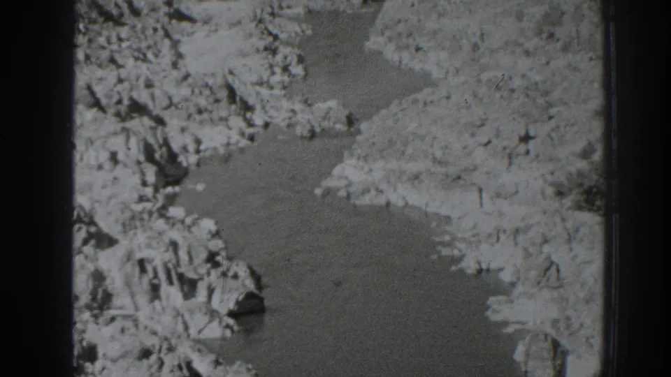 Home-movie still showing a shallow creek edge, rough stones, and light dirt banks in a rural Oroville landscape.