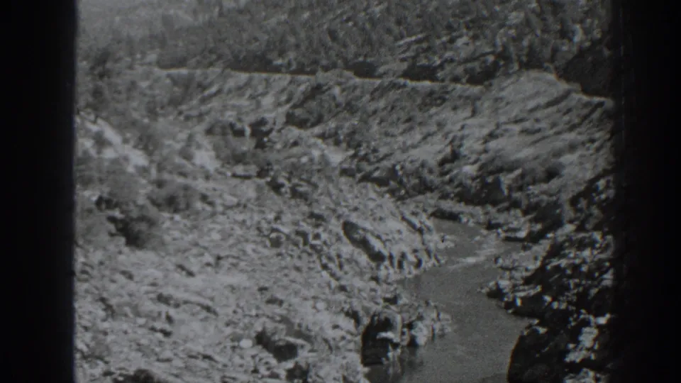 Soft black-and-white 8mm frame of dry Oroville-area rocks and a narrow stream channel, with visible film grain and dark edging.