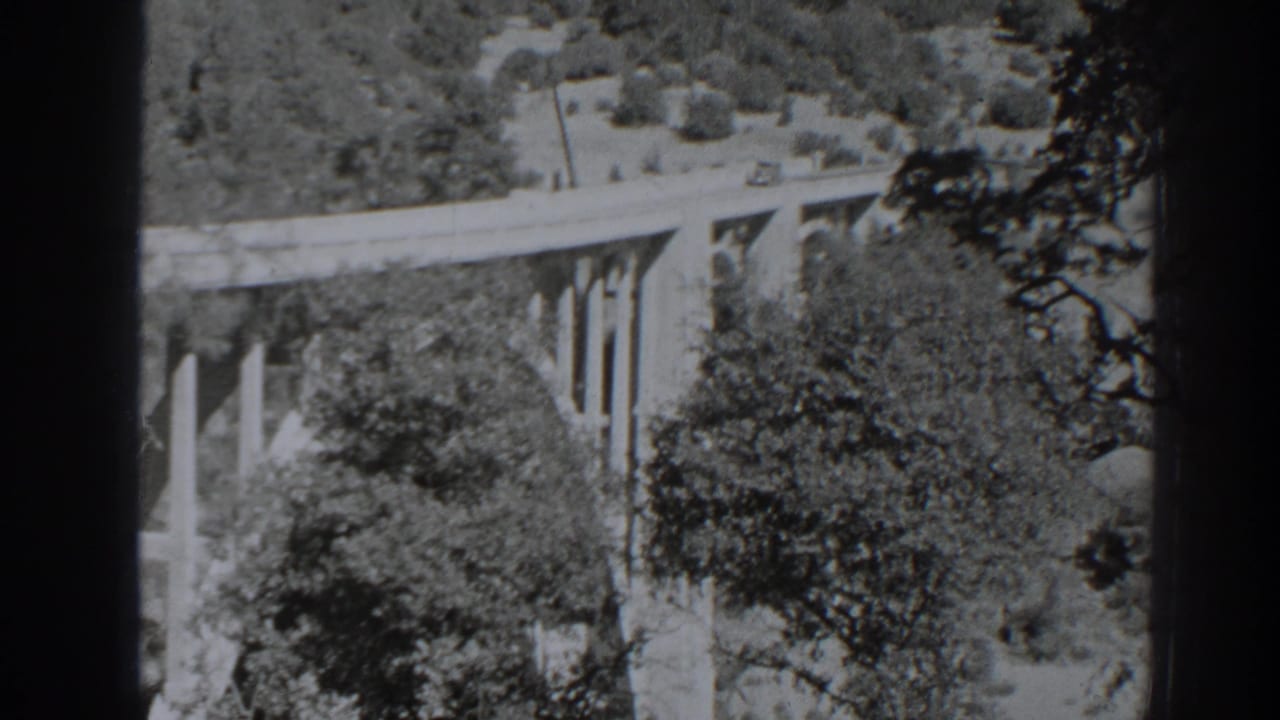 1938 Oroville-area 8mm frame showing a car on an elevated concrete bridge surrounded by wooded slopes.
