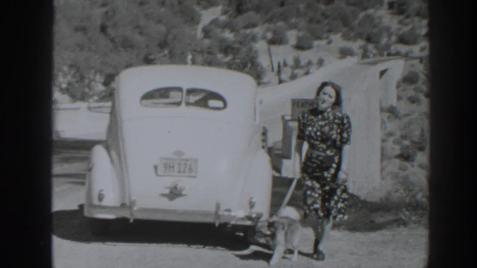 Woman in a patterned dress poses by a classic car and Feather River sign in the stated 1938 Oroville, California footage.