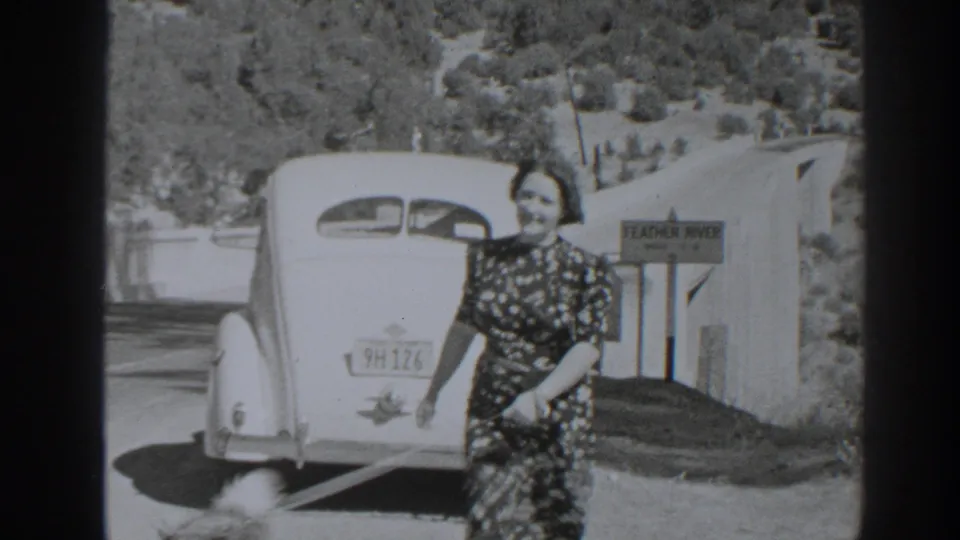 Woman with a small leashed dog moves beside the parked 1930s car near the Feather River sign in the Oroville footage.